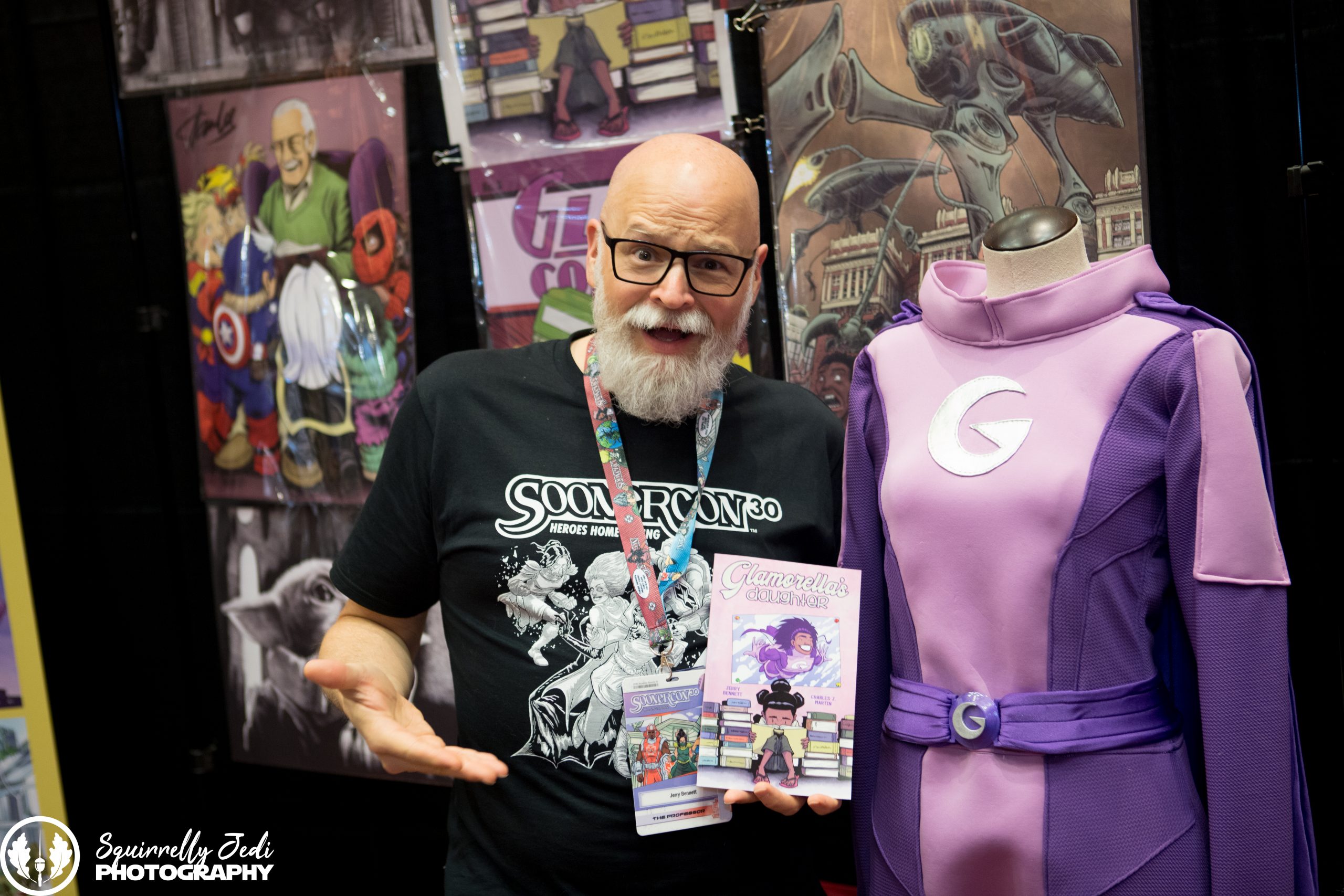 Artist Jerry Bennett gives a goofy style to the camera while holding his comic book Glamorella's Daughter and standing next to a Glamorella costume on a mannequin.